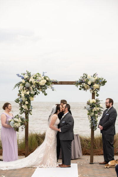 Bride and groom kissing under floral arch during outdoor wedding ceremony.