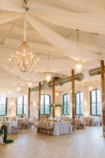 Elegant wedding reception setup in a spacious, rustic venue with chandeliers and draped ceilings.