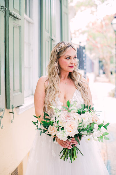 Bride in a white dress holding a bouquet, standing by a window.