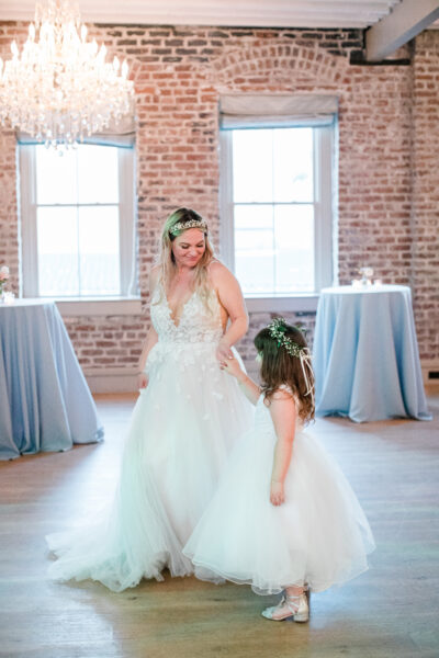 Bride in a white gown with a flower girl in a bright, rustic room.