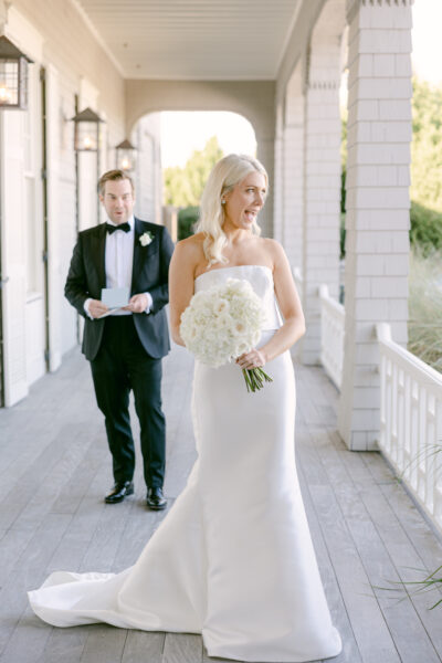 Bride in white gown holding bouquet with groom in tuxedo behind her on veranda.