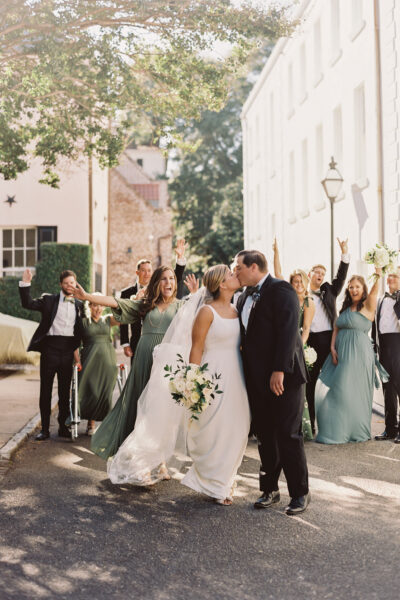 Bride and groom kiss as wedding party celebrates outdoors.
