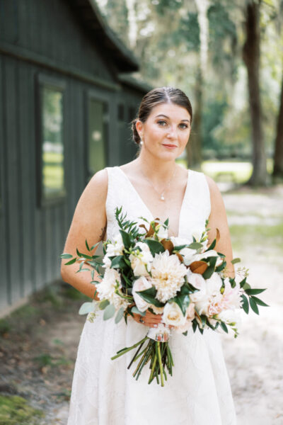 Bride in white dress holding a large floral bouquet outdoors.