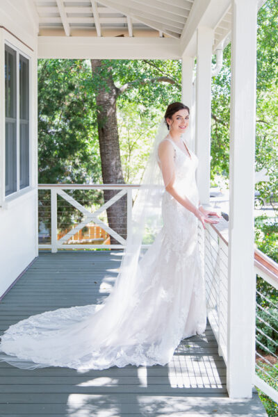 Bride in elegant wedding dress standing on porch with greenery outside.