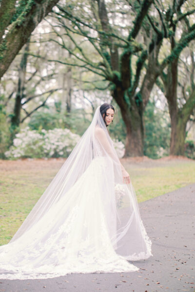 Bride in a long white gown and veil walking outdoors under blossoming trees.