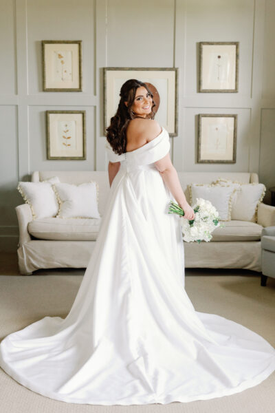 Bride in elegant off-shoulder white gown holding bouquet, standing in stylish living room.