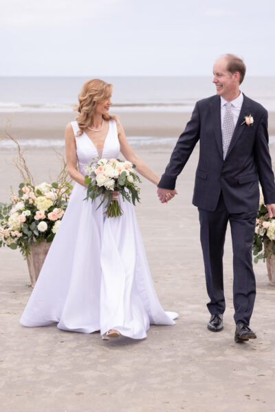 Newlyweds holding hands on a beach, bride in white gown and groom in black suit.