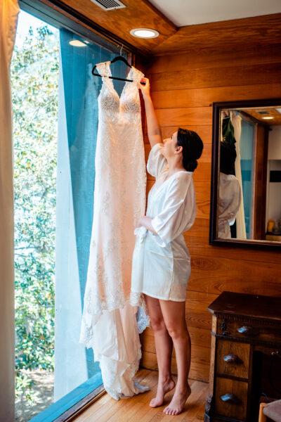 Woman in white robe reaching for a wedding dress hanging by a window.