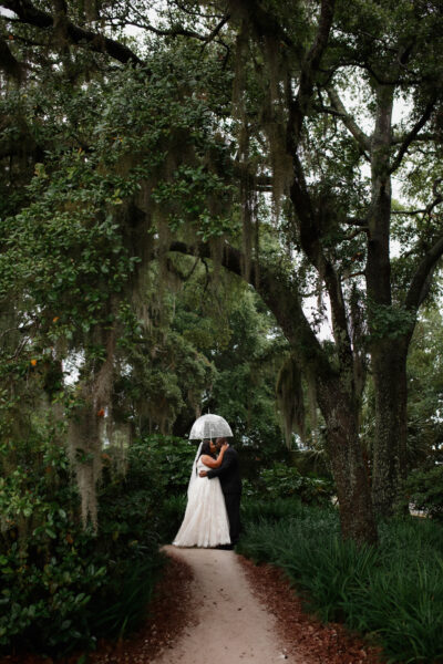 A couple shares a romantic embrace under large, leafy trees.