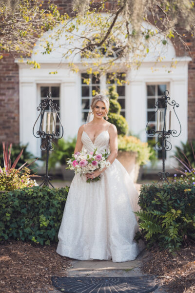 Bride in elegant gown holding bouquet, smiling outdoors.