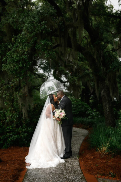 Bride and groom share a kiss under an umbrella in a lush forest.