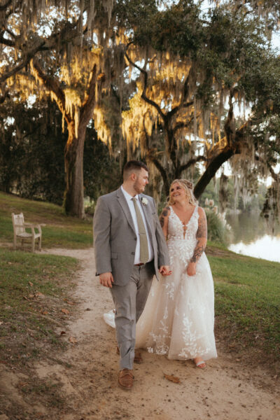 Newlywed couple walking hand in hand outdoors under moss-draped trees.