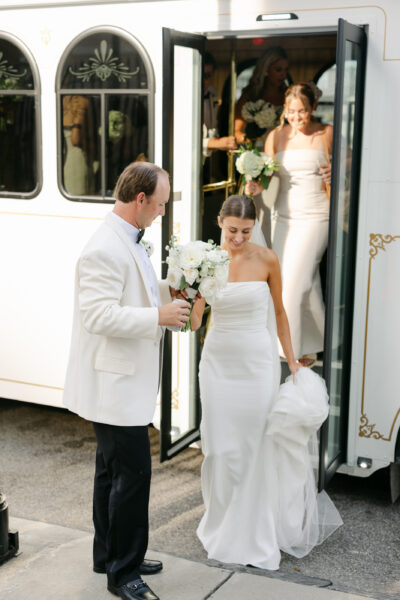 Bride in elegant white gown holding flowers, accompanied by a man in a white suit near a vehicle.