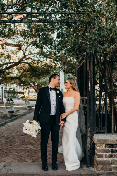 Newlyweds holding hands under a tree-lined path, sharing a joyful moment.