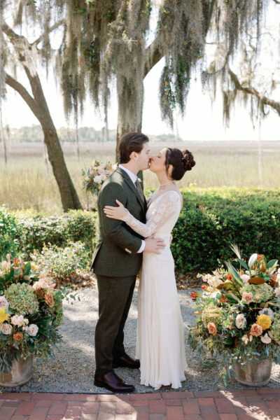 Bride and groom share a kiss surrounded by floral arrangements outdoors.