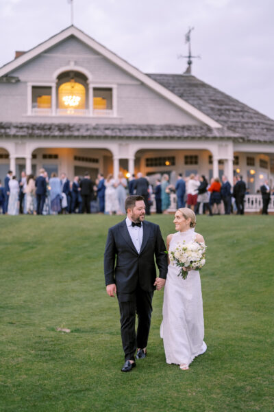 Bride and groom walking on grass with guests at a house in the background.