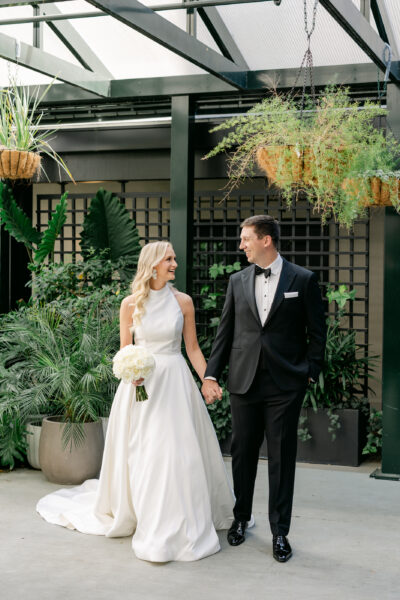 Bride and groom holding hands outdoors with greenery.