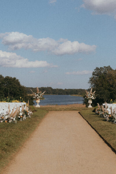 Outdoor wedding aisle with white chairs set beside a lake under a blue sky.