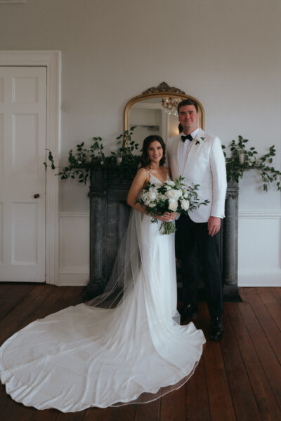 Bride and groom standing together indoors, bride in white gown and groom in white tuxedo.