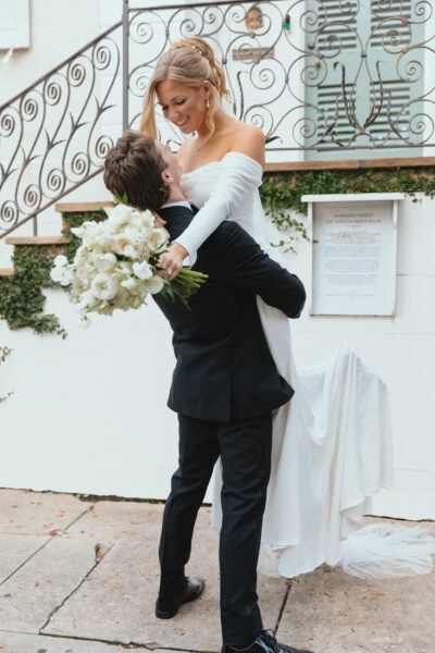 Groom lifts bride holding a bouquet outside a charming venue.