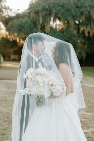 Newlyweds share a tender moment under a veil with a bouquet of white flowers.
