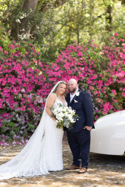 Bride and groom smiling in front of vibrant pink flowers on their wedding day.