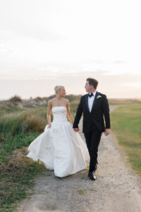Bride and groom walking hand in hand outdoors at sunset.