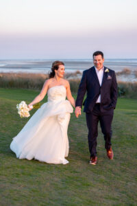 Newlywed couple holding hands and walking outdoors at sunset.