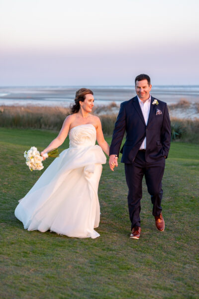 Newlywed couple holding hands and walking outdoors at sunset.