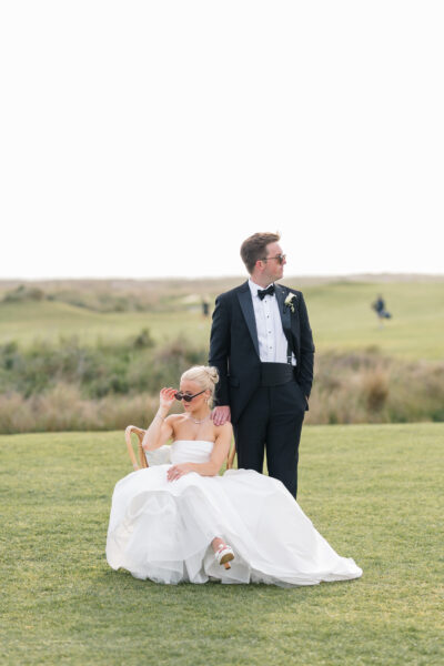 Bride sitting on the grass looking at groom standing nearby in a scenic outdoor setting.