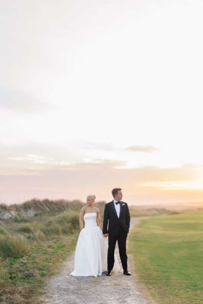 Bride and groom standing on grassy dunes at sunset.