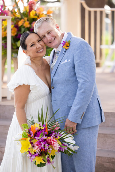 Newlywed couple smiling, holding a vibrant bouquet outdoors.